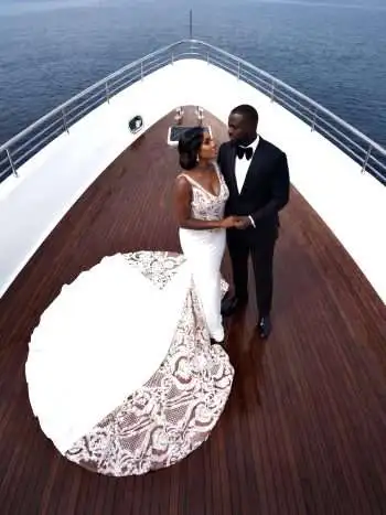 Bride and groom on a yacht deck, holding hands; she wears a white lace wedding gown with a long train, he in a black tuxedo.